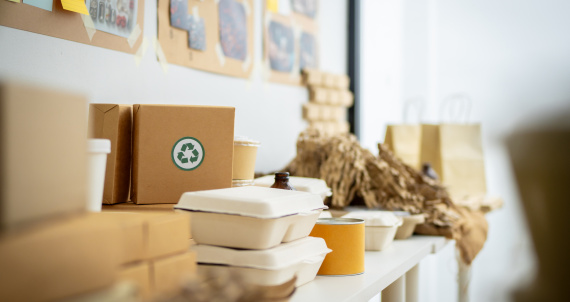 A stack of cardboard boxes with green recycling symbols on them. The boxes are piled on top of each other, and there are also some paper cups and bowls in the background