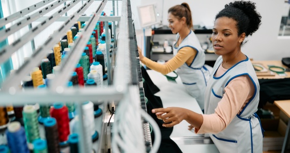 African American seamstress working at embroidery machine in a textile factory. Young black woman and her coworker working at embroidery machine at clothing factory.